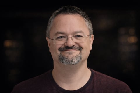 Middle-aged man with glasses and goatee wearing a dark maroon shirt, smiling at camera against a dark background