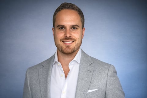 Professional headshot of a man with short brown hair wearing a gray blazer and white shirt against a blue background