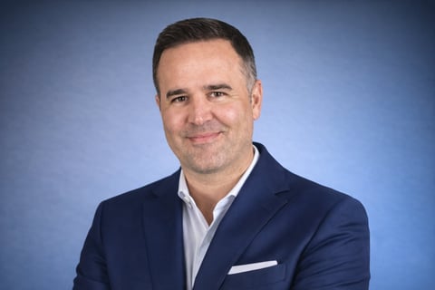 Professional headshot of a man wearing a dark blue blazer and white shirt against a blue background