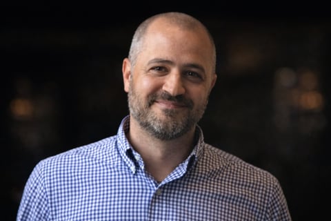Middle-aged man with gray beard and receding hairline smiling at camera while wearing a light blue checkered shirt against a dark blurred background