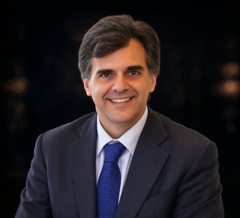 Professional headshot of a man in a dark navy suit and blue tie, smiling at the camera against a dark background