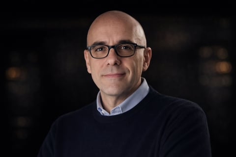 Bald man wearing glasses and dark sweater with collared shirt underneath, smiling at camera against dark blurred background