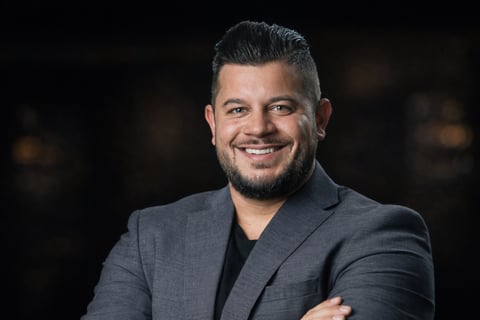 Professional headshot of a smiling man wearing a gray blazer and dark shirt against a blurred dark background