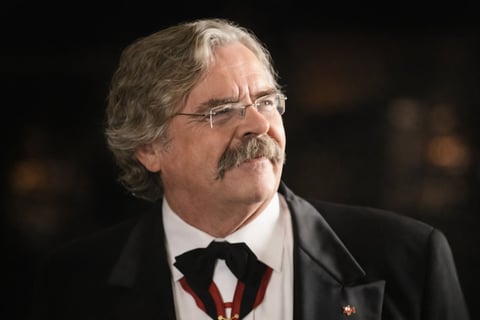 Middle-aged man with gray hair and mustache wearing glasses, tuxedo with black bow tie against dark background