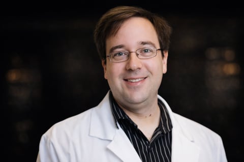 Man wearing glasses and white lab coat over striped shirt, smiling at camera against dark background