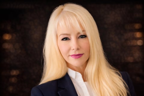 Professional headshot of a woman with long blonde hair, bangs, wearing a dark blazer and white shirt against a blurred dark background