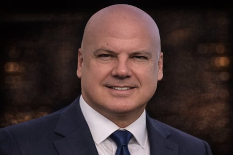 Bald man in dark suit and blue tie smiling at camera against blurred brown background
