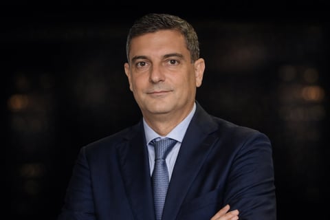 Professional headshot of a man in a dark navy blazer and blue tie with arms crossed against a dark background