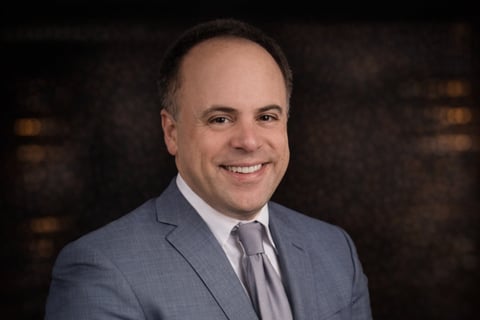 Professional headshot of a smiling middle-aged man wearing a blue blazer and gray tie against a blurred dark background